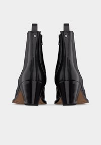 Ζευγάρι μαύρων γυαλιστερών δερμάτινων μποτάκια αστραγάλου με τακούνια μπλοκ και πίσω λουράκια, φαίνεται από πίσω πάνω σε λευκό φόντο.