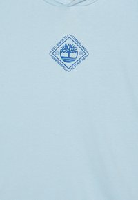 Svetlo modra kapuca s mehko teksturo. Ima modro grafično logotip v obliki diamanta z napisom "Timberland EST. SINCE 73."