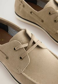 Pantofi din suede perforat bej, cu șireturi plate, ochiuri metalice și o talpă din cauciuc alb contrastant. Au un căptușeală din material striped.