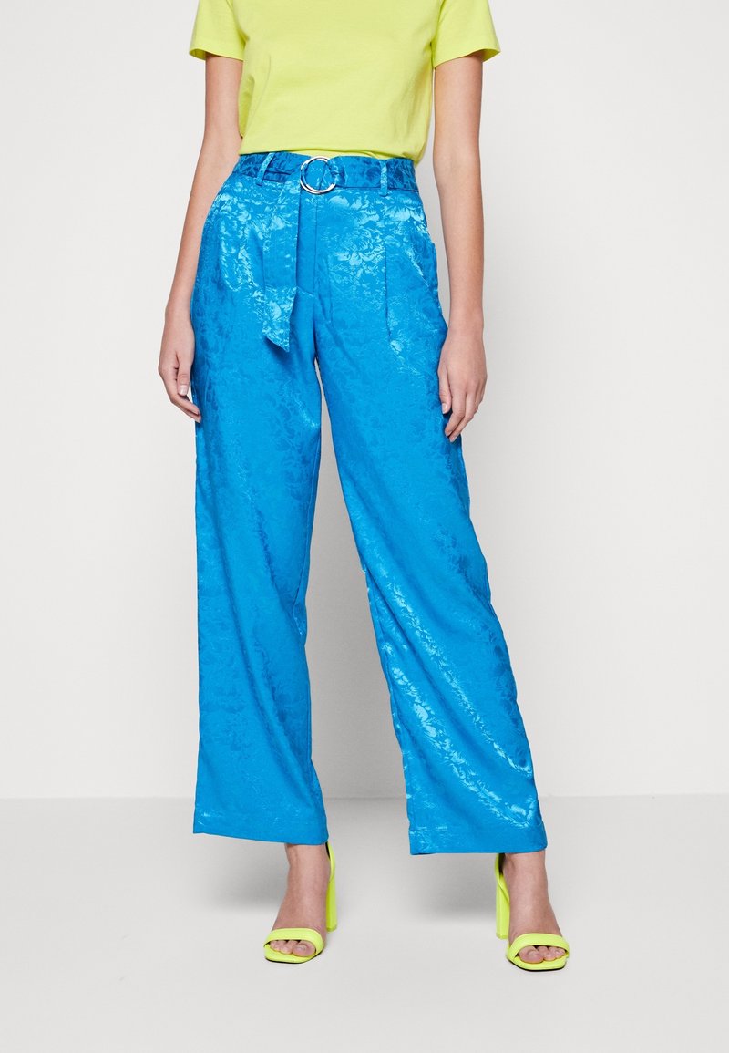 Femme portant un pantalon large taille haute bleu brillant avec ceinture, un haut vert citron et des sandales à talons hauts vert citron assorties.