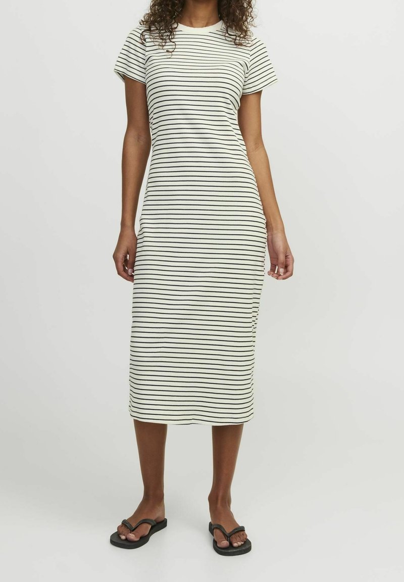 Robe midi rayée en crème et noir, en tissu côtelé. Caractéristiques : manches courtes et décolleté rond. Portée avec des tongs noires.