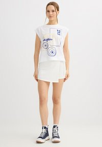 Joven mujer con camiseta sin mangas blanca con gráfico, falda blanca asimétrica, calcetines blancos y zapatillas altas azul marino, de pie sobre fondo liso.