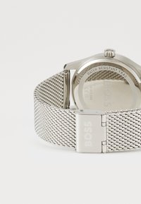 Reloj con correa de malla plateada y caja redonda. Cuenta con un cierre de marca y un fondo resistente al agua. Diseño minimalista con un acabado pulido.