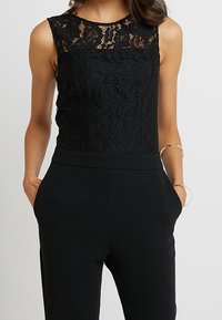 Top de encaje negro con un patrón floral, combinado con pantalones negros ajustados. El top tiene un escote redondo y un diseño sin mangas.