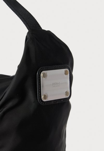 Gros plan d’un coin de sac en tissu noir avec une plaque d’étiquette métallique carrée fixée par quatre rivets, présentant un marquage discret "FPM Milano".