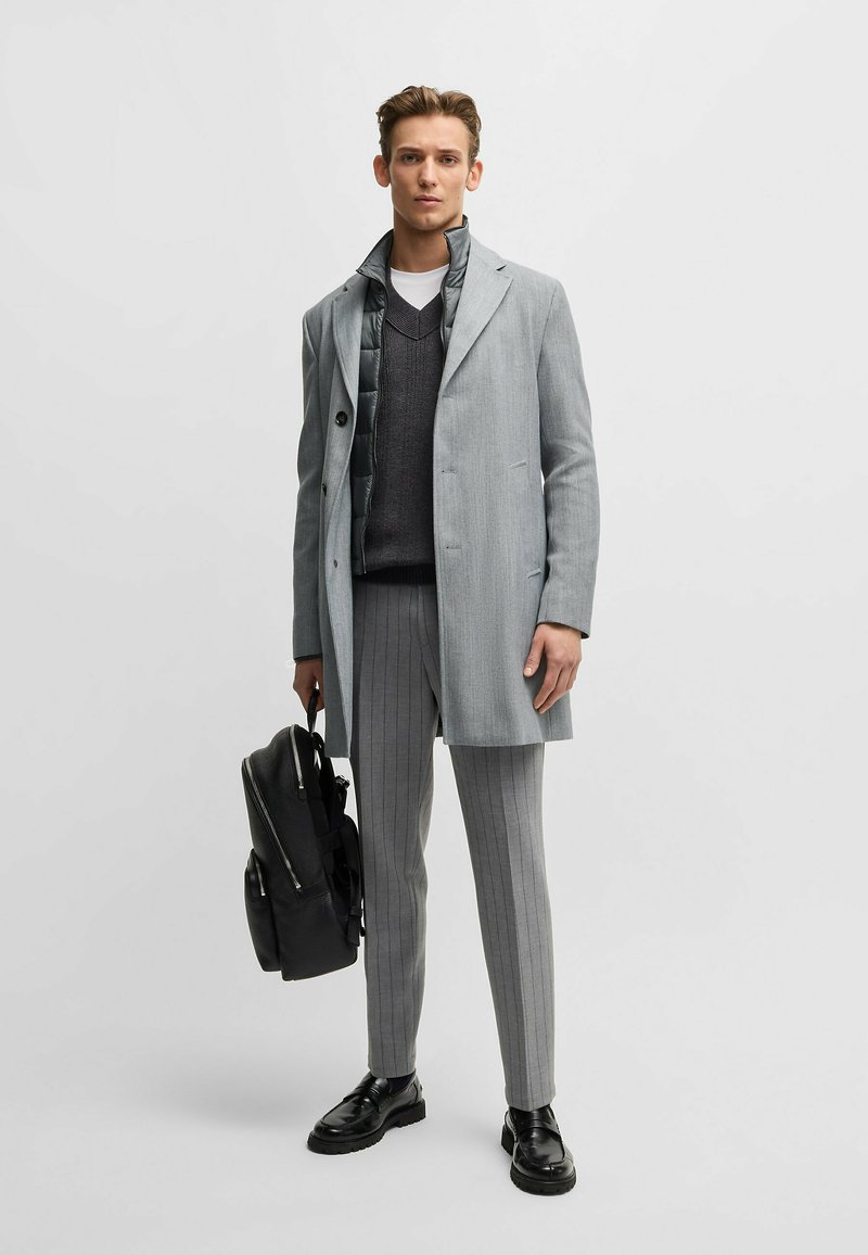 Ljusgrå kappa med slagkrage, lager på lager över en mörkgrå tröja och randiga byxor. Svart ryggsäck med dragkedjor och texturerad yta.