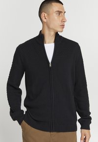 Sort strikket cardigan med fuld lynlås, tekstureret øverste del og ribbede manchetter. Har en høj krave og en glat underkrop.