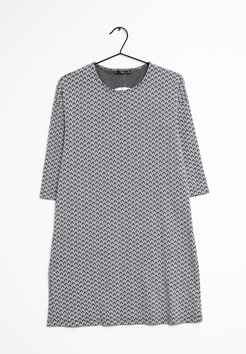 Robe à manches longues noire et blanche avec un motif en zigzag, suspendue à un cintre en métal noir contre un fond blanc.