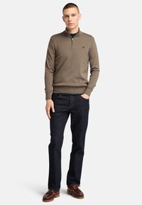 Brązowy sweter z zamkiem, wykonany z miękkiej dzianiny, połączony z ciemnoniebieskimi dżinsami i brązowymi butami w stylu boat shoes, stojący na jednolitym tle.