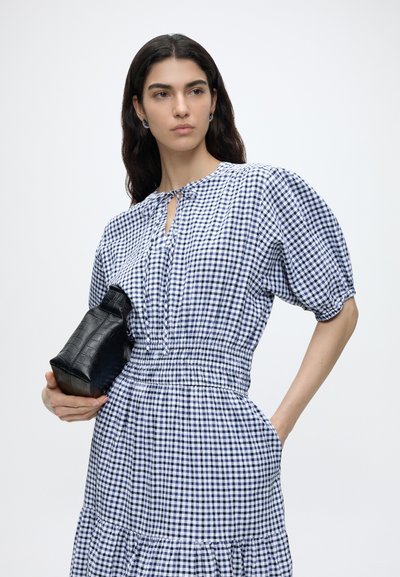 Femme aux longs cheveux foncés portant une robe à carreaux bleus et blancs, tenant une pochette noire texturée sur un fond uni.