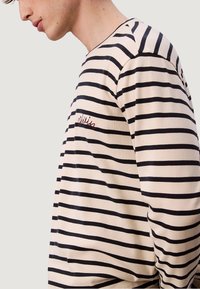 Mladý člověk v béžovém a černém pruhovaném tričku s dlouhým rukávem, na hrudi s malým červeným vyšitým textem, otočený vlevo.