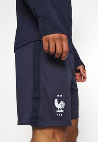Osoba ubrana w granatowe sportowe szorty z białym emblematem koguta i dwiema gwiazdkami, połączone z pasującą bluzą z długim rękawem.