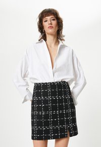 Chemise blanche à manches lâches associée à une jupe noire avec un motif à carreaux blancs et une fente sur le côté.