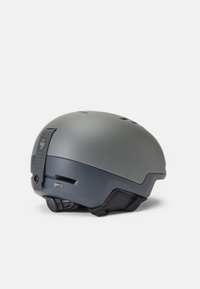 Casco gris con un acabado mate liso, forma aerodinámica, ranuras de ventilación y un detalle de correa ajustable en la parte posterior.