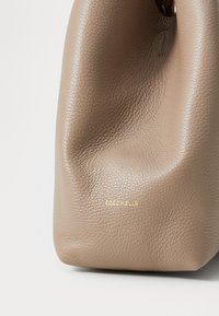 Borsa a mano in pelle beige con superficie testurizzata, caratterizzata da un logo dorato "Coccinelle" e una forma arrotondata e morbida.