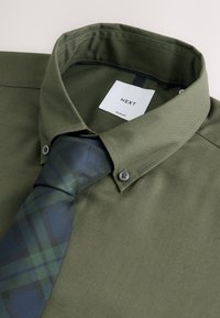Camisa ajustada de color verde oliva con cuello doblado y cierre de botones, combinada con una corbata de cuadros en verde oscuro y azul marino. Tela suave y texturizada.