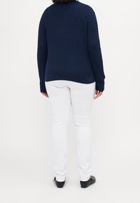Donkerblauwe gebreide trui met een gestructureerd patroon, geribbelde zoom en manchetten, gecombineerd met witte broek. Zijaanzicht van de persoon die de outfit draagt.