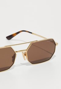 Ochelari de soare octagonali cu nuanțe de maro, având un cadru din metal auriu. Prezintă un model de țestoasă pe brățară și un logo pe lentilă.