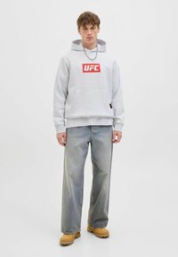 Junger Mann in einem grauen UFC-Hoodie, hellblauen weit geschnittenen Jeans, braunen Stiefeln und einer silbernen Kette, die vor einem schlichten weißen Hintergrund steht.