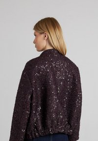 Una donna bionda di profilo a sinistra indossa una giacca di paillettes viola scuro con colletto alto e orlo elastico su uno sfondo grigio chiaro.