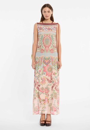 Frau in einem bodenlangen, ärmellosen Kleid mit mehrfarbigen Blumenmustern, die aufrecht vor einem weißen Hintergrund steht.