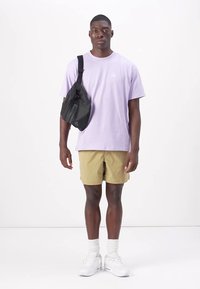 Lys lilla bomulls t-skjorte med liten hvit logo, kombinert med tan shorts og hvite sneakers. Svart crossbody-veske hengende over venstre skulder.