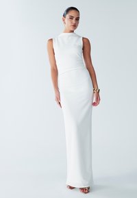 Robe maxi blanche sans manches en tissu lisse et extensible. Elle présente un col haut et une silhouette ajustée, avec un léger éclat.