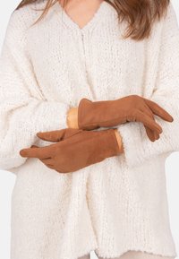 Gants en suede marron doux avec une texture lisse, un design ajusté et des détails subtils, portés sur un pull en maille épaisse blanc.