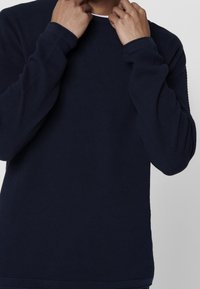 Laivastonsininen teksturoitu neulepaita, jossa on pitkät hihat, pyöreä pääntie ja resorit hihansuissa. Kangas on kudottu kuvioitu lisäämään ulottuvuutta.