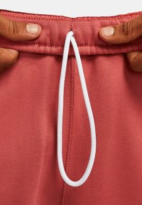 Cintura de pantalones cortos de tela roja con un cordón blanco. La cintura presenta un acabado de costura fina y una textura suave. Manos sostienen la cintura.