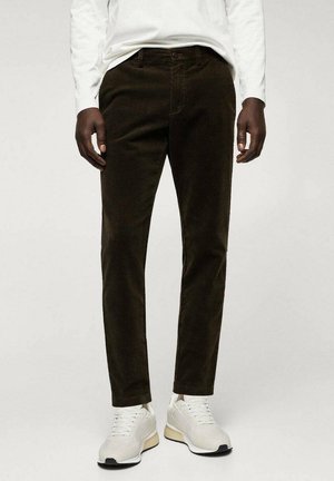 Man draagt donkerbruine corduroy broek, witte sneakers en een wit overhemd met lange mouwen, staande tegen een effen lichte achtergrond.