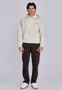Felpa beige con cappuccio e tasca frontale, abbinata a pantaloni marrone scuro con logo ricamato, indossata con scarpe da ginnastica bianche.