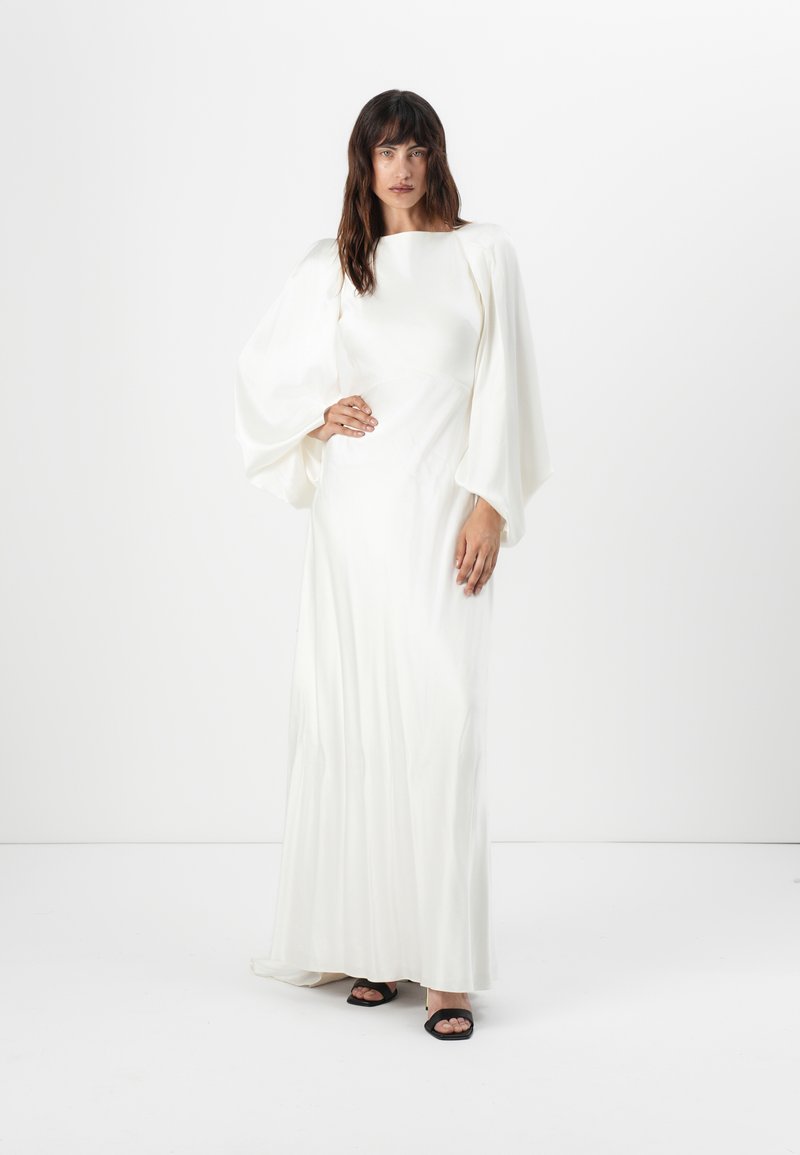 Robe blanche à manches longues avec un décolleté structuré et un tissu fluide, présentant des manches bouffantes et une silhouette droite. Chaussures noires à bout ouvert.