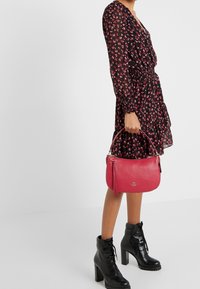 Sac à main en cuir rouge de forme arrondie, avec des ferrures argentées et un accent de pompon. Porté à l'avant-bras, associé à une robe fleurie et des bottes noires.