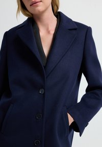 Marineblauer Wollmantel mit Reverskragen, einreihigem Design mit fünf Knöpfen und Seitentaschen. Weiche Textur und taillierte Passform.