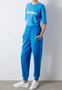 Blue cotton t-shirt with "publishers." text and matching blue joggers with side pockets and elastic cuffs. White sneakers complete the outfit.