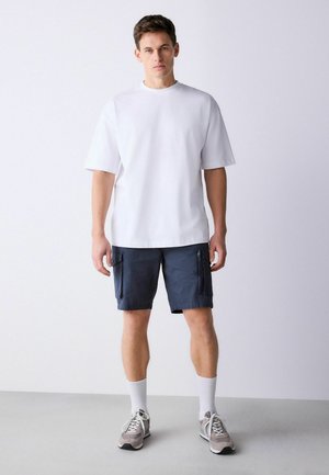Homme debout portant un t-shirt blanc uni oversize, un short cargo bleu marine, des chaussettes blanches montantes et des baskets grises sur un sol gris.