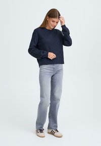 Sötétkék kötött pulóver, laza szabással, kerek nyakkal és bordázott textúrával; világosszürke farmernadrággal és krémszínű, fekete csíkkal ellátott tornacipővel párosítva.