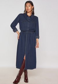 Abito in denim con maniche lunghe, chiusura con bottoni, due tasche sul petto, vita elastica e spacco anteriore. Colore blu scuro, con una texture liscia.