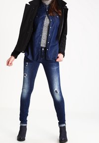 Vaqueros de mezclilla de corte ajustado, lavado azul medio, con detalles desgastados, combinados con una camisa de manga larga azul marino y una chaqueta negra. Botines negros.