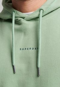 Grön huvtröja med slät textur, med dragsnören med metallspetsar och "SUPERDRY"-logotypen i marinblått tryckt på framsidan.