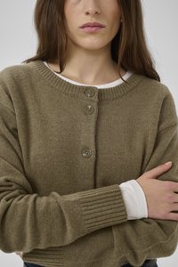 Frau trägt eine olivgrüne, mit Knöpfen versehene Strickjacke über einem weißen Langarmshirt und hat die Arme verschränkt.