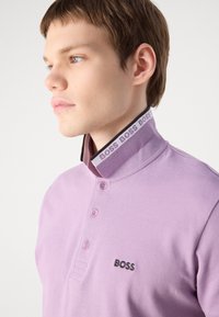Ljubičasta polo majica s crnim kragnom. Ima dugmad i izvezeno "BOSS" logo na lijevoj strani grudi. Glatka tekstura, uski kroj.