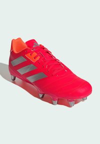 Botín de fútbol de un vibrante color rosa con acentos rojos, parte superior de material sintético, logo de tres rayas en gris y seis tacos para tracción.