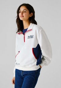 Biely fleecový pulóver s námorníckymi a červenými akcentmi, ktorý má štvrtinový zips, logo na hrudníku a bočné vrecká. Textúrovaná tkanina.