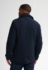 Cappotto in misto lana blu navale con colletto alto, spalle strutturate e polsini con bottoni. Include tasche laterali e una vestibilità rifinita.