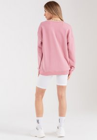 Różowy, oversize'owy sweter z prążkowanymi mankietami, stylizowany z białymi, teksturowanymi szortami rowerowymi oraz białymi sneakersami. Widok z tyłu pokazujący luźny krój i szczegóły dekoltu.