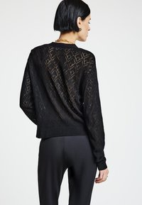 Maglione nero a rete aperta con motivo geometrico, polsini e orlo a costine. Presenta una vestibilità rilassata e maniche lunghe. Indossato sopra pantaloni neri.