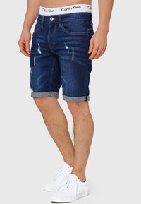 Traper šorts u tamnoplavoj boji s izlizanim mrljama, srolanim rubom, dizajnom s pet džepova i Calvin Klein logom na elastičnom pojasu. Upareni s bijelim tenisicama.