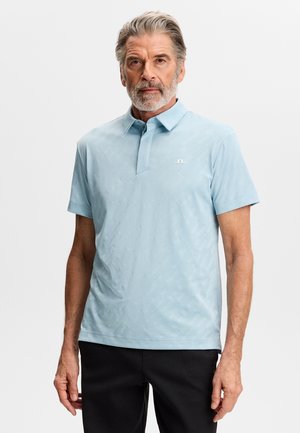 Uomo anziano con capelli e barba grigi che indossa una polo a maniche corte azzurro chiaro e pantaloni neri, in piedi davanti a uno sfondo semplice.